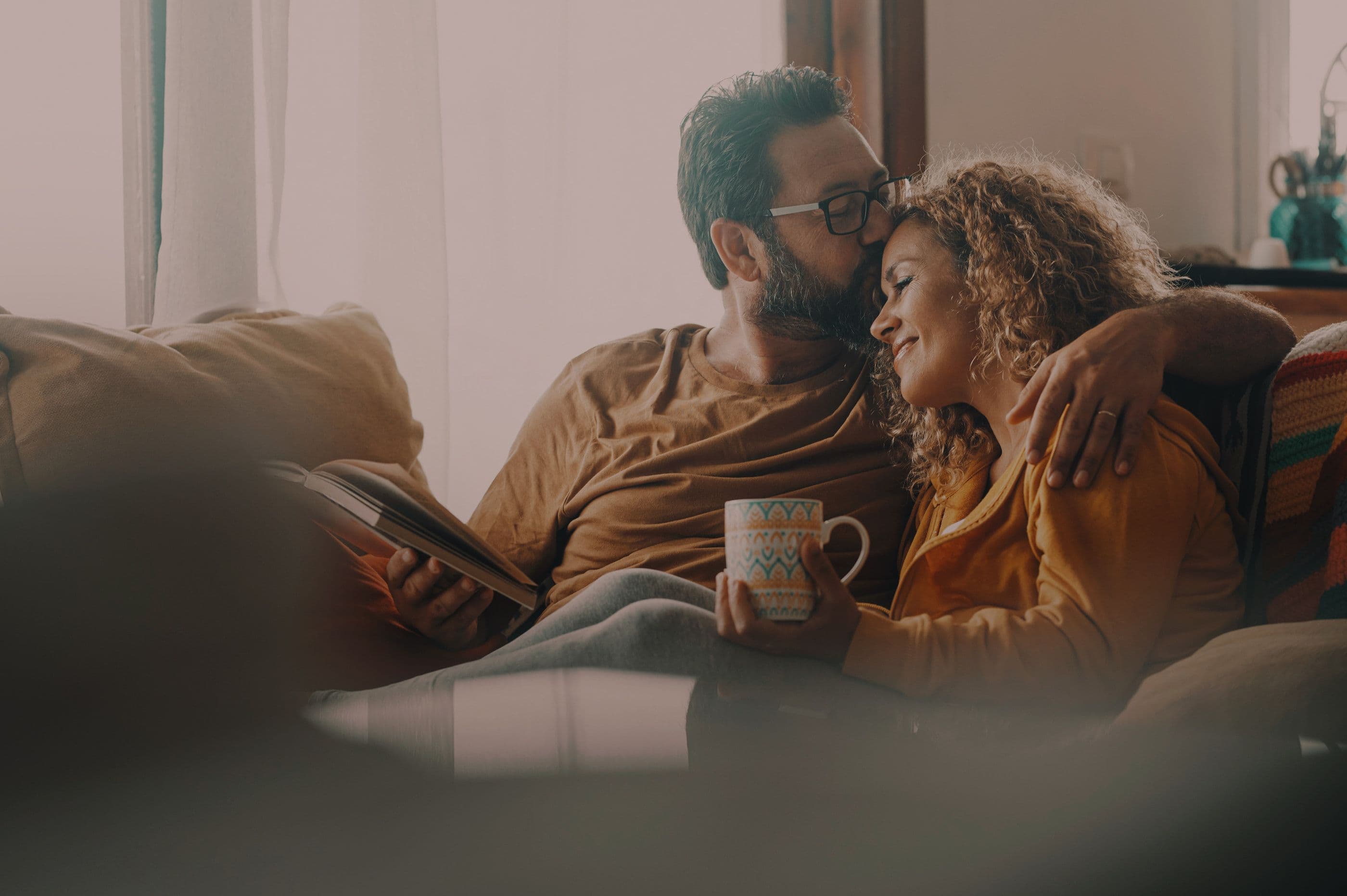 Happy middle-aged couple reading and drinking from a mug. Man kisses woman on forehead.